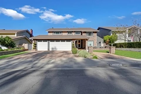 Two-story suburban home with beige and brown exterior, two-car garage, and manicured lawn under clear blue sky